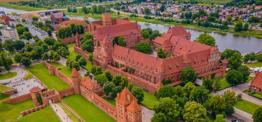 The red-brick Gothic walls and towers of Malbork Castle reflected in the Nogat river at sunset.